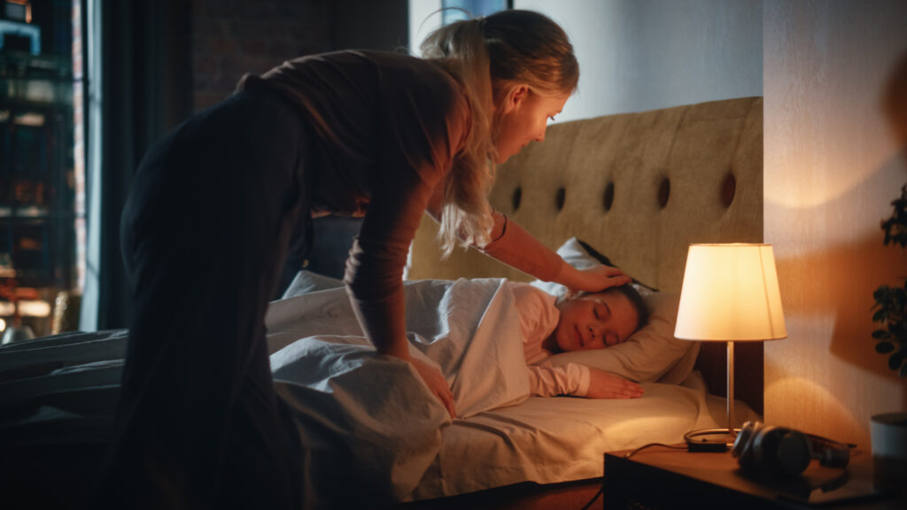 Parent tucking a child into bed at night with a warm bedside lamp on.